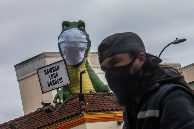 Un hombre que lleva una máscara recomendada contra el coronavirus  pasa junto a un dinosaurio en la cima del Odditorium Ripleys Believe It or Not con una máscara facial junto a un letrero que alienta a las personas a lavarse las manos en Hollywood.