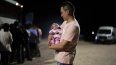Un migrante carga a su hija en brazos tras cruzar la frontera, mientras otros inmigrantes abordan un autobus.&nbsp;