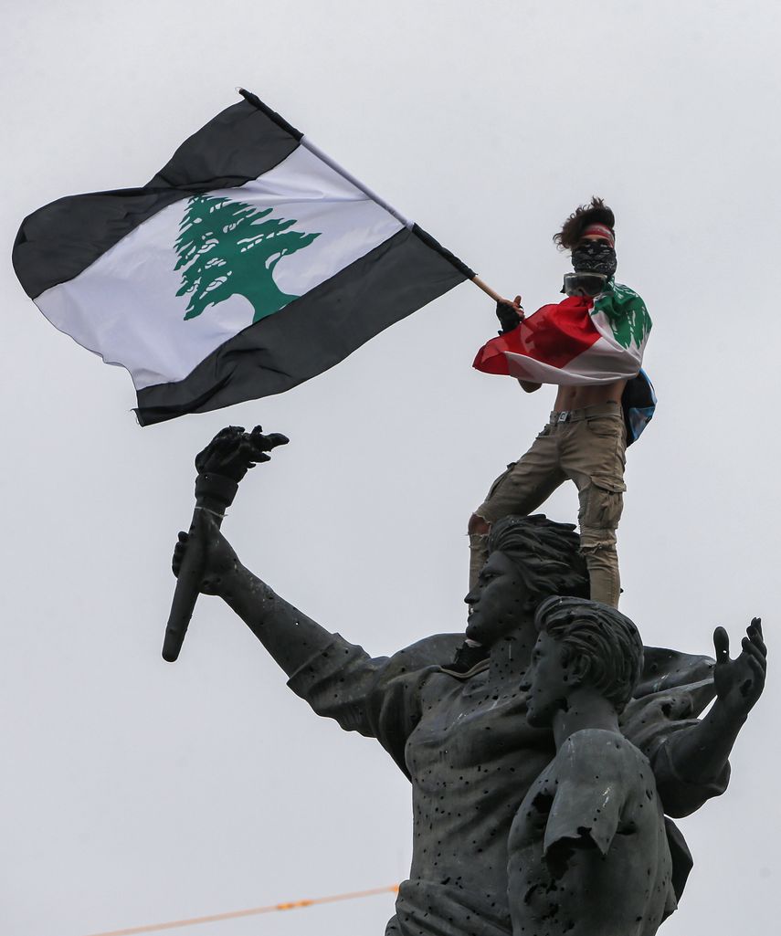 Un manifestante con una bandera libanesa con rayas negras se encuentra sobre la estatua de los m&aacute;rtires en la Plaza de los M&aacute;rtires en el centro de la capital del L&iacute;bano, Beirut, durante una manifestaci&oacute;n contra el gobierno el 1 de septiembre de 2020.