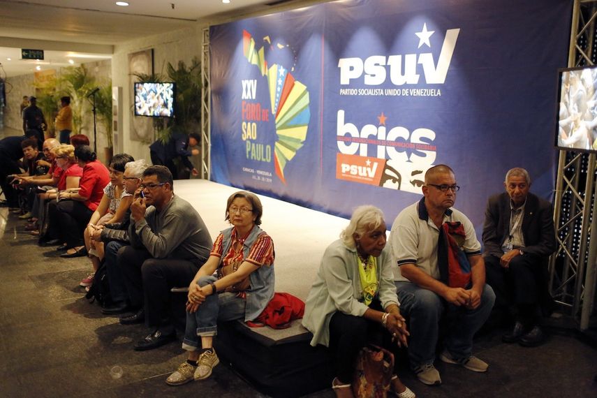 Varios participantes permanecen sentados cerca de un carteles del Foro de Sao Paulo y del Partido Socialista Unido de Venezuela durante la primera jornada de esa reunión en Venezuela, el viernes 26 de julio de 2019.&nbsp;