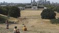 Gente sentada en pasto seco por el sol en el Parque de Greenwich, con el Museo Marítimo y el distrito financiero de Canary Wharf al fondo, en Londres, el domingo 17 de julio de 2022.&nbsp;