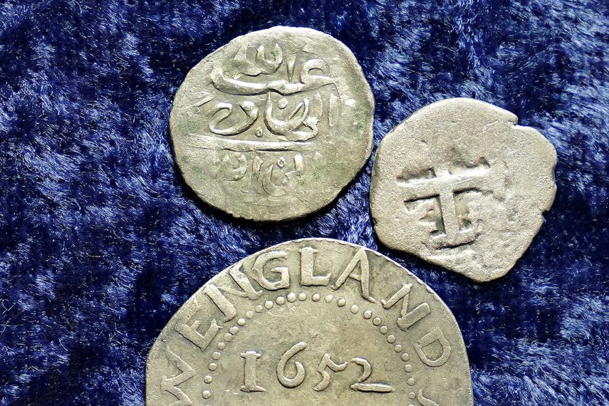 Una moneda de plata árabe del siglo XVII, en la parte superior, que según las investigaciones se acuñó en 1693 en Yemen, descansa cerca de un chelín de roble acuñado en 1652 por la colonia de la bahía de Massachusetts, abajo, y una moneda española de medio real de 1727, a la derecha, sobre una mesa , en Warwick, RI, el jueves 11 de marzo de 2021.&nbsp;