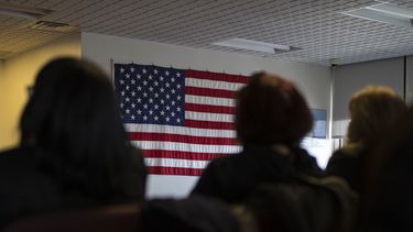 Varias personas esperan en una habitación frente a la bandera de los EEUU.