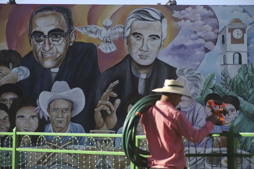 Un jardinero con sus herramientas camina frente a un mural con las imágenes del difunto arzobispo Óscar Romero y el padre Rutilio Grande el viernes 21 de enero de 2022 en la Plaza de Los Mártires, en El Paisnal, El Salvador.&nbsp;&nbsp;