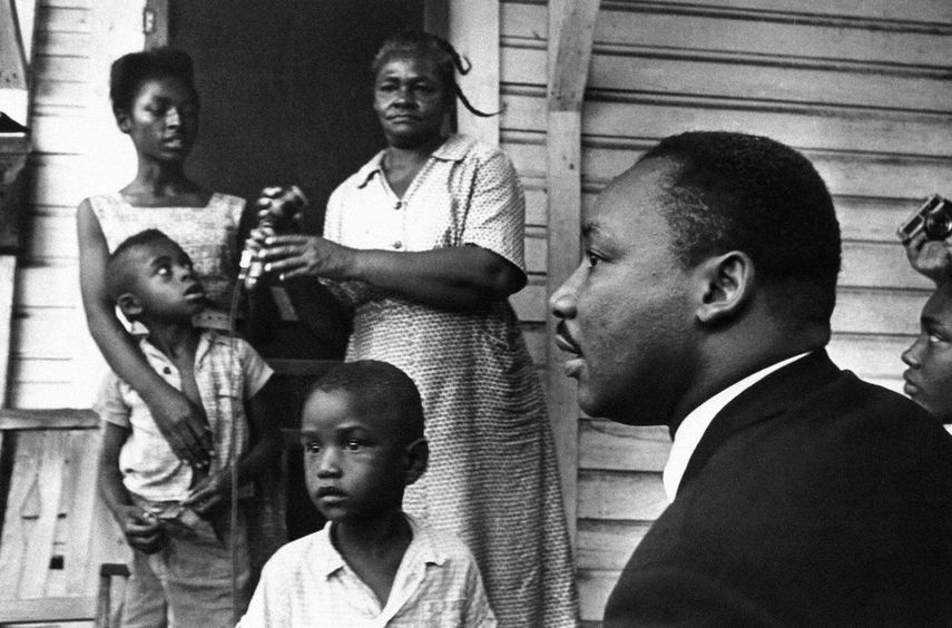 El Dr. Martin Luther King Jr., a la derecha, conversa con afroamericanos de Greenwood, Mississippi, en el porche de su casa el 21 de julio de 1964, durante su campaña puerta a puerta, diciéndoles que se registren para votar y apoyen a su partido Demócrata por la Libertad de Mississippi.
