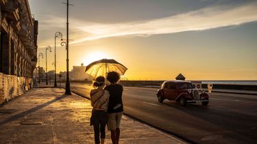 Una pareja con mascarillas para protegerse del nuevo coronavirus sostiene un paraguas mientras camina por el malecón al atardecer en La Habana, Cuba, el lunes 31 de agosto de 2020. Las autoridades de La Habana dispusieron prorrogar la noche del viernes un paquete de medidas restrictivas que incluyen el toque de queda nocturno en la capital mientras tratan de controlar un rebrote de COVID-19.