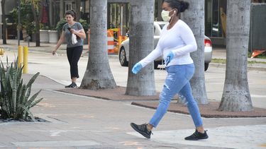 Una mujer camina por una calle de Miami, protegida por una mascarilla, mientras otra lleva la suya en la mano.