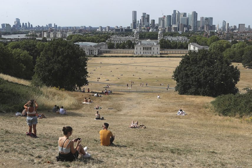 Gente sentada en pasto seco por el sol en el Parque de Greenwich, con el Museo Marítimo y el distrito financiero de Canary Wharf al fondo, en Londres, el domingo 17 de julio de 2022.&nbsp;