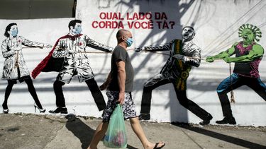 Un hombre que porta una mascarilla camina el viernes 19 de junio de 2020 frente a un mural en Sao Paulo, Brasil, que describe una lucha entre trabajadores de salud por un lado y el presidente Jair Bolsonaro y un personaje que representa al coronavirus por el otro, con un mensaje en portugués que dice: ¿De qué lado de la cuerda estás tú?&nbsp;