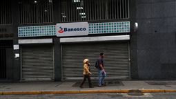 Dos personas caminan frente a un negocio cerrado este lunes, en el marco de la suspensión de actividades laborales y escolares, en Caracas