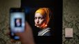 Un visitante toma una foto con su teléfono móvil de una versión diseñada con inteligencia artificial por el creador digital con sede en Berlín Julian van Dieken (C) inspirado en la pintura de Johannes Vermeer La joven de la perla en el museo Mauritshuis en La Haya el 9 de marzo de 2023. Artistas rechazan programas de inteligencia artificial que imitan sus diseños sin darles crédito.&nbsp;