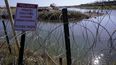Migrantes cruzan el río Bravo a Estados Unidos desde México detrás de un hilo de cuchillas y un cartel que advierte que cruzar es peligroso e ilegal, el miércoles 3 de enero de 2024 en Eagle Pass, Texas.&nbsp;