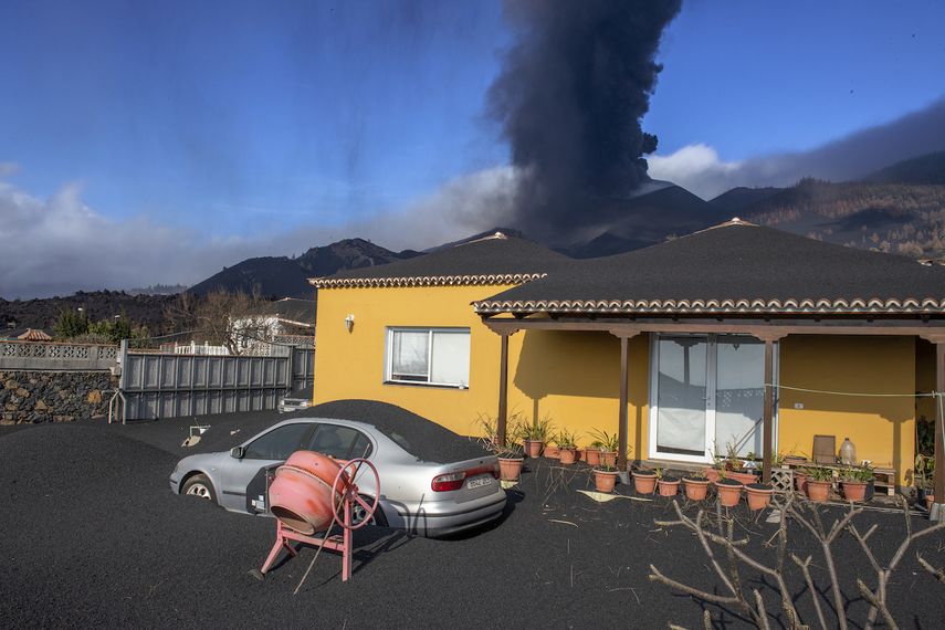 Una capa de ceniza volcánica cubre una casa, un automóvil y sus alrededores mientras en el fondo un volcán sigue en erupción en la isla canaria de La Palma, España, el lunes 4 de octubre de 2021.&nbsp;