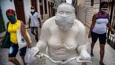   Peatones con mascarilla para protegerse del coronavirus pasan junto a una escultura que alguien adornó con una mascarilla para promover la distancia social, en La Habana, Cuba.   