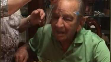 Abuelo de 100 años intenta soplar las velas en su cumpleaños con un protector facial puesta prodcuto de la pandemia del COVID-19