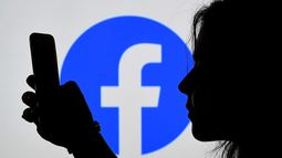 Una usuaria de Facebook navega en su celular. En el fonde el logo de la red social.