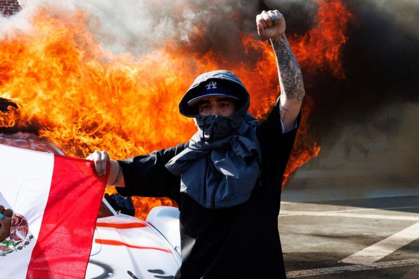 Un manifestante que participa en los disturbios en Los Ángeles levanta el puño mientras sostiene una bandera mexicana frente a un vehículo de Waymo que fue incendiado durante una manifestación tras las operaciones federales de inmigración en Los Ángeles.