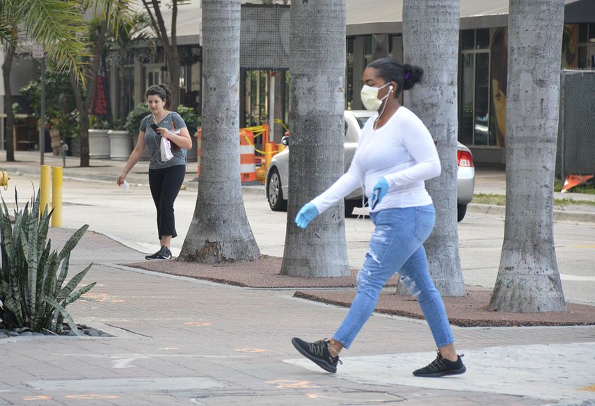 Una mujer camina por una calle de Miami, protegida por una mascarilla, mientras otra lleva la suya en la mano.