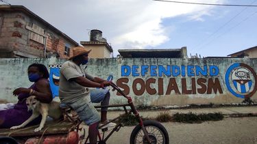 La gente monta un triciclo cerca de un grafiti que dice Defendiendo el Socialismo en La Habana,