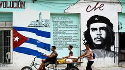 Varias personas transportan comida en un triciclo que pasa por una pared pintada con el glaseado cubano y un retrato del argentino Ernesto Che Guevara en una calle de La Habana el 27 de junio de 2023.