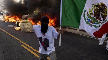 Un manifestante sostiene una máscara suave que imita al presidente de Estados Unidos, Donald Trump, y una bandera mexicana frente a autos Waymo en llamas mientras los manifestantes se enfrentan con las fuerzas del orden cerca del edificio federal durante una protesta en respuesta a las operaciones federales de inmigración en Los Ángeles, California, el 8 de junio de 2025.&nbsp; &nbsp;