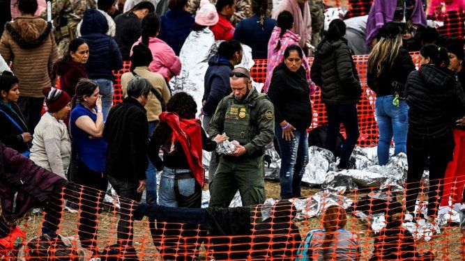Oficial de Aduanas y Protección Fronteriza de Estados Unidos (Centro) entrega comida a inmigrantes que esperan ser procesados en un centro de tránsito de la Patrulla Fronteriza después de cruzar la frontera desde México en Eagle Pass, Texas, el 22 de diciembre de 2023.