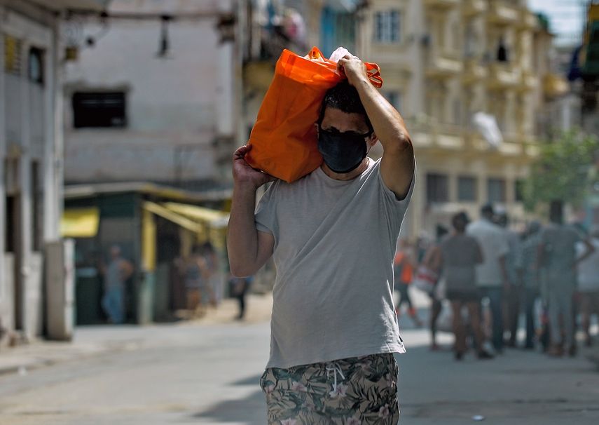 Un hombre sostiene una bolsa con comida en una calle en La Habana. Al fondo, un grupo de personas espera en una fila.