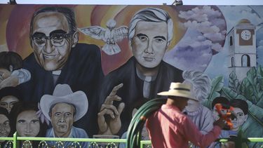 Un jardinero con sus herramientas camina frente a un mural con las imágenes del difunto arzobispo Óscar Romero y el padre Rutilio Grande el viernes 21 de enero de 2022 en la Plaza de Los Mártires, en El Paisnal, El Salvador.&nbsp;&nbsp;