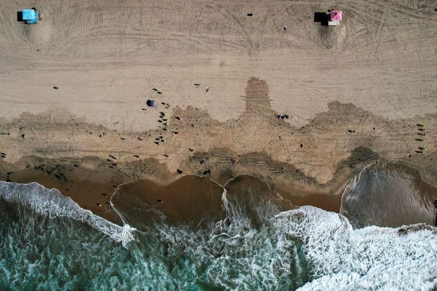 Esta fotografía tomada con un dron muestra desde las alturas a bañistas mientras trabajadores con trajes protectores continúan limpiando las partes contaminadas con crudo en la playa de la localidad de Huntington Beach, California.