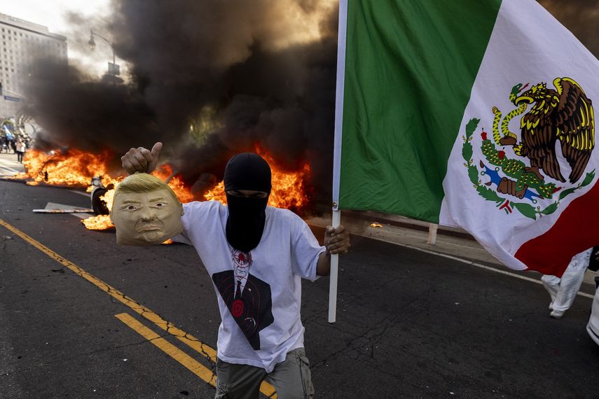 Un manifestante sostiene una máscara suave que imita a Donald Trump y una bandera mexicana frente a autos Waymo en llamas mientras los manifestantes se enfrentan con las fuerzas del orden cerca del edificio federal durante una protesta en respuesta a las operaciones federales de inmigración en Los Ángeles, California, el 8 de junio de 2025.&nbsp; &nbsp;