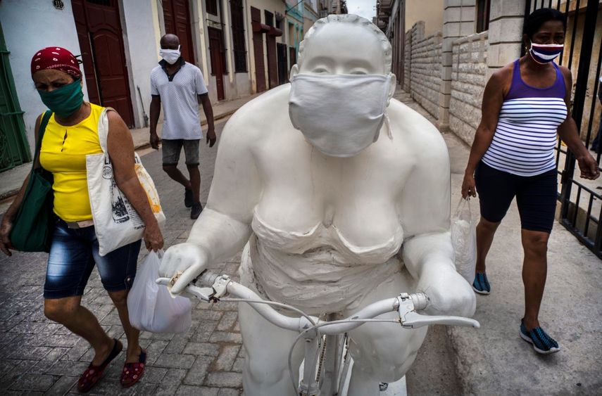   Peatones con mascarilla para protegerse del coronavirus pasan junto a una escultura que alguien adornó con una mascarilla para promover la distancia social, en La Habana, Cuba.   