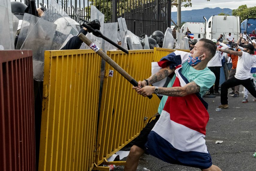 Un manifestante envuelto en una bandera costarricense y con un tubo de metal se enfrenta a la policía antidisturbios durante una protesta contra la moción del gobierno de aumentar los impuestos para llegar a un acuerdo crediticio con el Fondo Monetario Internacional (FMI), frente a la casa presidencial en San José. el 12 de octubre de 2020.
