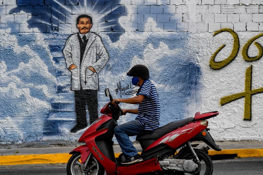 Un hombre con mascarillas pasa su motocicleta frente a un mural que representa al médico venezolano José Gregorio Hernández en una calle de Caracas, el 27 de abril de 2021. La Iglesia católica beatificará al médico venezolano José Gregorio Hernández el 30 de abril en Caracas.