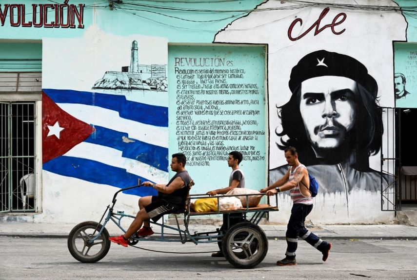 Varias personas transportan comida en un triciclo que pasa por una pared pintada con el glaseado cubano y un retrato del argentino Ernesto Che Guevara en una calle de La Habana el 27 de junio de 2023.