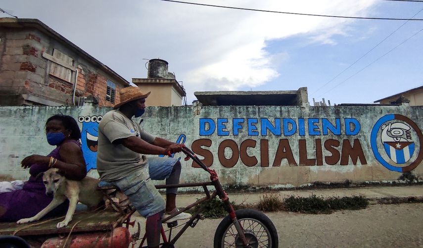 La gente monta un triciclo cerca de un grafiti que dice Defendiendo el Socialismo en La Habana,