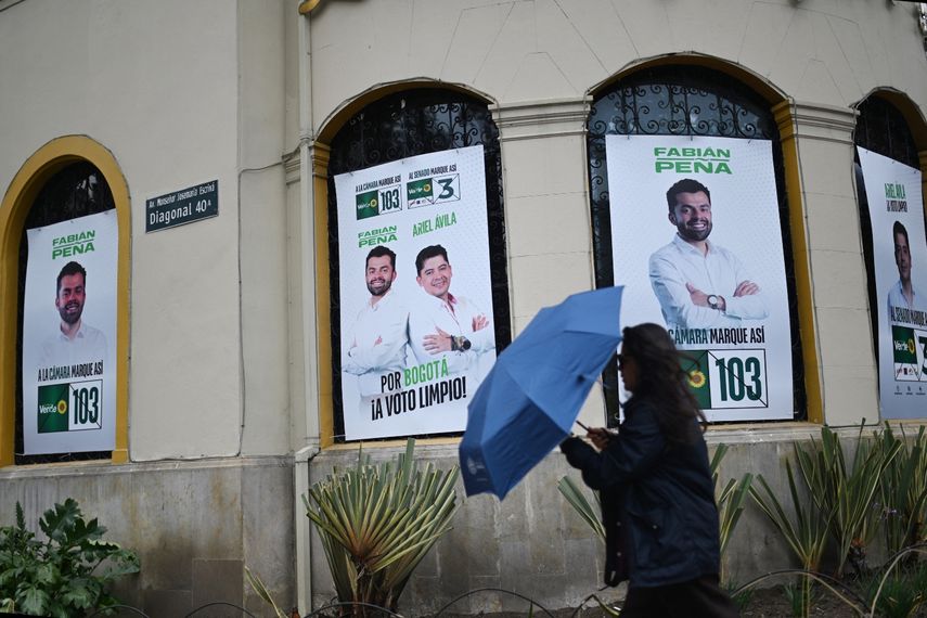 Una mujer camina con un paraguas junto a carteles de la campaña electoral de los candidatos a la cámara y al Senado de la Alianza Verde, Fabián Peña y Ariel Ávila, en Bogotá, el 3 de marzo de 2026. Colombia celebrará elecciones legislativas el 8 de marzo