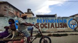 La gente monta un triciclo cerca de un grafiti que dice Defendiendo el Socialismo en La Habana,