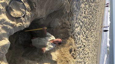 En esta fotografía proporcionada por la localidad de Kill Devil Hills, Carolina del Norte, David Elder, supervisor de rescate en el océano, permanece de pie el domingo 15 de mayo de 2022 en un agujero en la playa.