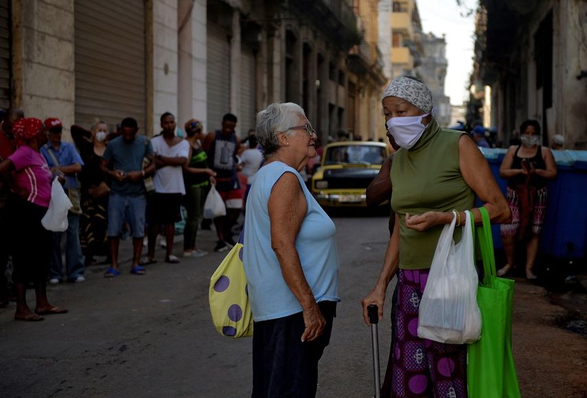 Dos mujeres de avanzada edad, una de ellas con una mascarilla, conversan junto a una fila de personas que esperan para adquirir suministros en una calle en La Habana el 24 de marzo de 2020, en medio de la&nbsp;emergencia social por la&nbsp;propagaci&oacute;n&nbsp;del nuevo coronavirus&nbsp;en Cuba.