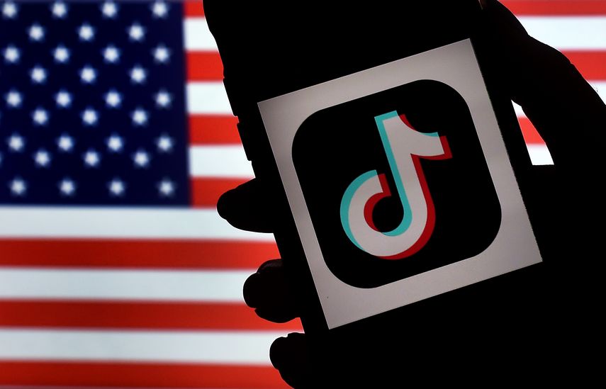 El logo de Tiktok en la pantalla de un tel&eacute;fono celular y la bandera de EEUU de fondo.