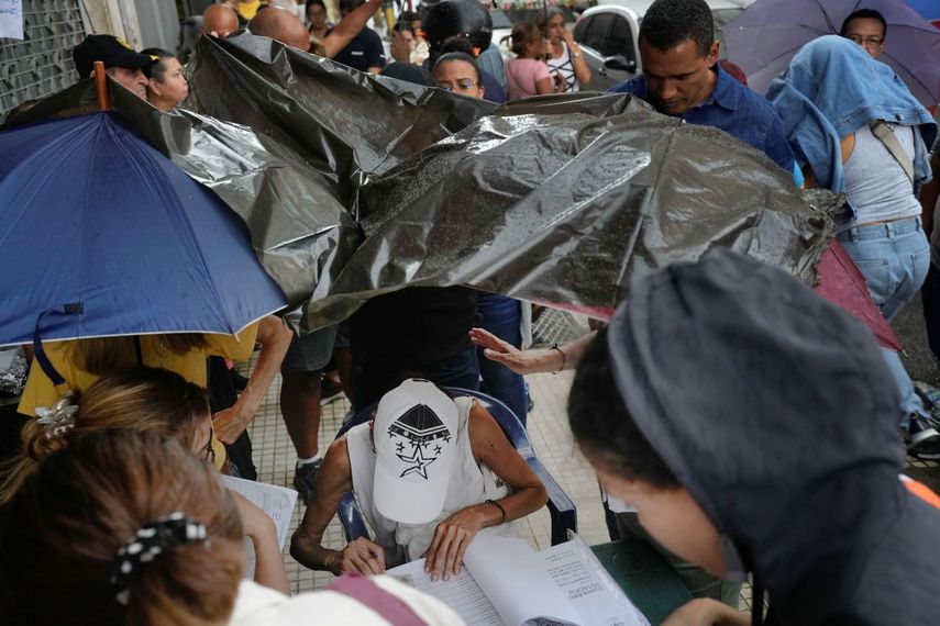 Una mujer, cubierta de la copiosa lluvia por una bolsa de plásticos, busca en una lista los nombres de los votantes para que puedan sufragar durante las elecciones primarias de la oposición en Caracas, Venezuela, el domingo 22 de octubre de 2023.&nbsp;