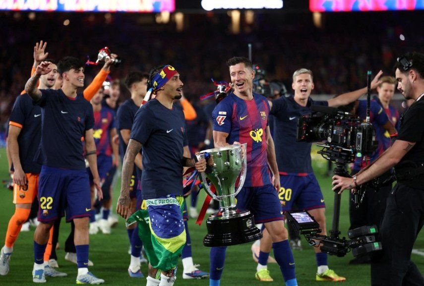 El delantero brasileño del Barcelona, Raphinha (número 11), y el delantero polaco del Barcelona, Robert Lewandowski (número 9), sostienen el trofeo mientras los jugadores del Barcelona celebran la victoria en la Liga española 2025-2026&nbsp;
