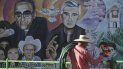 Un jardinero con sus herramientas camina frente a un mural con las imágenes del difunto arzobispo Óscar Romero y el padre Rutilio Grande el viernes 21 de enero de 2022 en la Plaza de Los Mártires, en El Paisnal, El Salvador.  