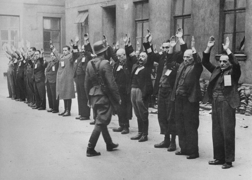 Un hombre nazi de las SS inspecciona a un grupo de trabajadores judíos en abril de 1943 en el gueto de Varsovia. En noviembre de 1940, los alemanes establecieron el gueto de Varsovia. La población judía que aún vivía fuera fue llevada al interior del área especial, y a los polacos que vivían dentro de los límites designados del gueto se les ordenó que se mudaran. El 15 de noviembre a ningún judío se le permitió salir del recinto judío. En el verano de 1942, unos 300.000 judíos fueron deportados a Treblinka. Cuando los informes de asesinatos en masa en el centro de exterminio se filtraron al gueto, un grupo superviviente, en su mayoría jóvenes, formó una organización llamada Z.O.B. (Zydowska Organizacja Bojowa, Organización de Lucha Judía) llamando al pueblo judío a resistir. El 19 de abril de 1943 comenzó el levantamiento del gueto de Varsovia después de que las tropas y la policía alemanas entraran en el gueto para deportar a sus habitantes supervivientes. Setecientos cincuenta combatientes lucharon durante casi un mes. Pero el 16 de mayo de 1943 la revuelta terminó. Los alemanes habían aplastado lentamente la resistencia. De los más de 56.000 judíos capturados, unos 7.000 fueron fusilados y el resto fueron deportados a centros de exterminio o campos de concentración. 
