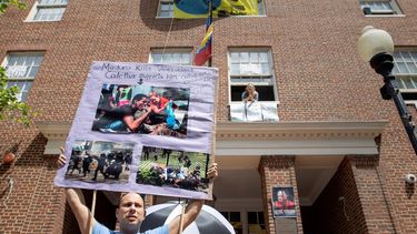 Desde la ventana una de las personas que ocupa la embajada de Venezuela en Washington, mientras un joven sostiene una pancartas denunciando la dictadura de Maduro.&nbsp;
