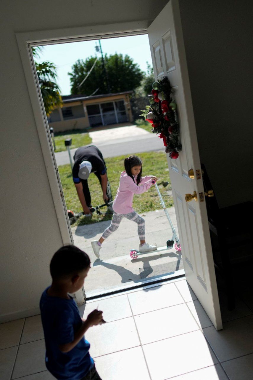 Alexa, de 7 años, juega con una patineta que recibió como regalo de Navidad mientras su padre, Alexis Llanos, se prepara para arreglar el jardín, y su hermano Alexis juega dentro de la casa a la que se mudaron en octubre de 2023.