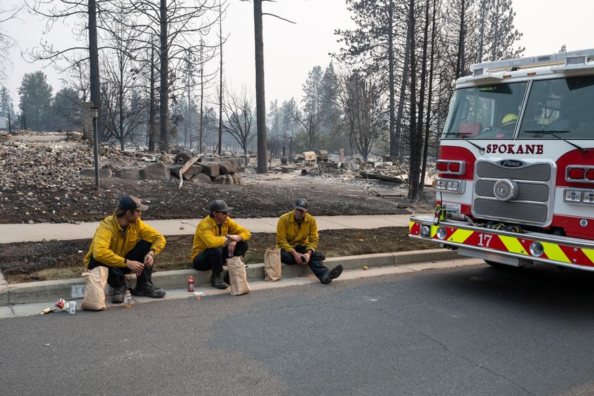 Tres miembros del Departamento de Bomberos de Spokane están sentados en el bordillo frente a viviendas calcinadas en el barrio de Balboa, en Spokane (Washington), el 2 de agosto de 2026