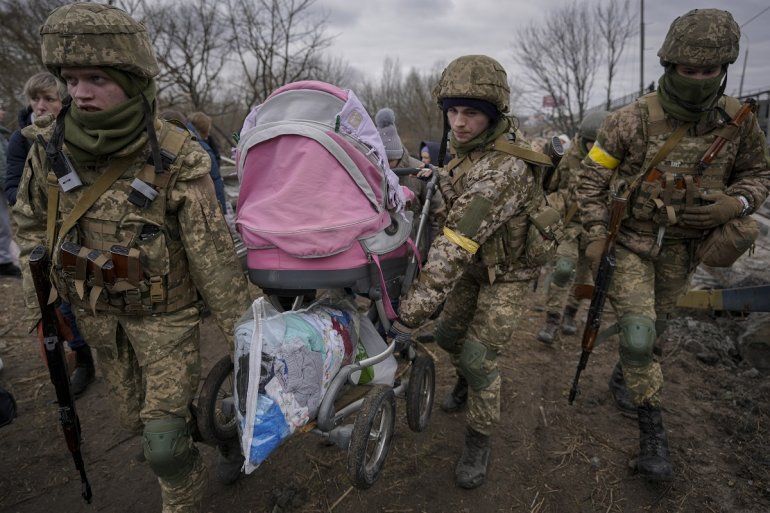 Militares ucranianos cargan un cochecito de bebé después de cruzar el río Irpin por un camino improvisado debajo de un puente que fue destruido por un ataque aéreo ruso, mientras ayudan a las personas que huyen de la ciudad de Irpin, Ucrania, el sábado 5 de marzo de 2022. 