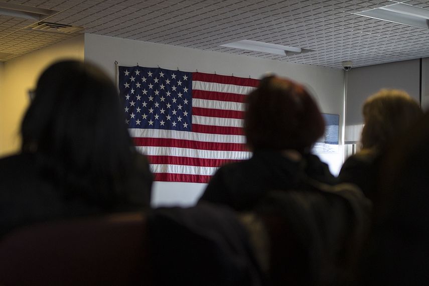 Varias personas esperan en una habitación frente a la bandera de los EEUU.