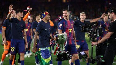 El delantero brasileño del Barcelona, Raphinha (número 11), y el delantero polaco del Barcelona, Robert Lewandowski (número 9), sostienen el trofeo mientras los jugadores del Barcelona celebran la victoria en la Liga española 2025-2026&nbsp;